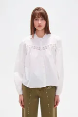 Camisa de algodón color blanco, con cuello mao, cierre frontal con botones y detalle de encaje en el pecho. Presenta mangas largas abullonadas con puños elásticos y un corte holgado.