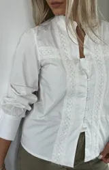 Camisa blanca de corte clásico con detalles de encaje en el frente y mangas largas con puños abotonados.