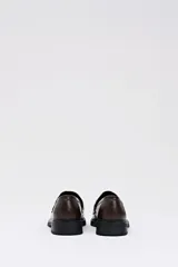 Mocasín tipo penny loafer de eco-piel color marrón chocolate, con suela negra gruesa y detalle de pelo con estampado de vaca en blanco y marrón en el empeine.