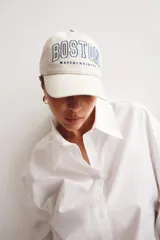 Gorra de sarga color crema con bordado azul marino de la palabra "BOSTON" y debajo "MASSACHUSETTS". Tiene botón en la parte superior, cinta antitranspirante y correa ajustable en la parte trasera.