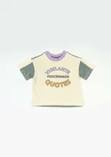 Remera color crema con cuello redondo y mangas cortas de color verde y lila. Estampado frontal con la frase "Vigilante Peacemaker Quotes" en letras color lila y naranja.