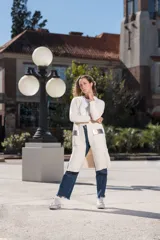 Saco largo color crudo, con cuello redondo, bolsillos frontales con apliques de cuero plateado y mangas largas.