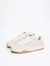 Zapatillas Tommy Jeans de corte bajo, color gris claro, confeccionadas en ante con interior de poliéster y suela de goma color caramelo. Presentan cordones blancos, logo de la marca en la lengüeta y etiqueta de Tommy Jeans en el lateral.