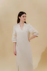 Vestido largo color beige, confeccionado en lino, con escote en V con volados y apliques de puntilla en las mangas. Posee bolsillos delanteros y tajos laterales.
