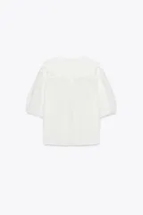 Camisa blanca de cuello bobo con bordados, escote pico con lazada, manga corta abullonada con elástico y cierre frontal con botones.