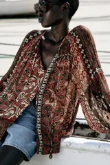 Blusa marrón con estampado de cachemir y detalles de hilo metalizado. Tiene cuello redondo con cordón ajustable y abalorios, escote en pico con nido de abeja, mangas largas con elástico en los puños y cierre frontal con botones forrados.