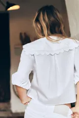 Camisa blanca de popelín con cuello bobo bordado y lazada. Tiene mangas abullonadas por debajo del codo con elástico y pinzas en la espalda. El cierre frontal es con lazadas.