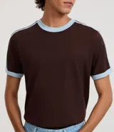 Remera de algodón de corte regular, color blanco hueso, con cuello redondo y ribete negro. Presenta un diseño de doble raya negra en las mangas y ribetes negros en el cuello y los puños de las mangas.