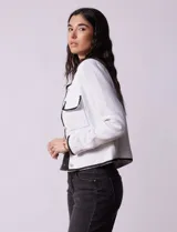 Chaqueta de tweed blanca con ribete negro, escote a la base, abertura central con botones, bolsillos delanteros y puños con aplique de botones perlados.