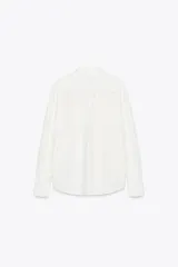 Camisa blanca de algodón con cuello solapa, manga larga con puños abotonados, bolsillo de parche en el pecho, bajo redondeado y cierre frontal con botones.