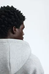 Campera con capucha y cierre frontal, color gris melange, de tejido efecto neopreno. Presenta un corte holgado (boxy) con hombros caídos y bolsillos delanteros abiertos.