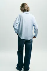 Camisa celeste de cuello solapa y manga larga con pliegues. Bajo asimétrico y cierre frontal con botones.