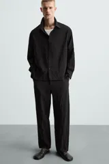 Pantalón negro de corte relajado (relaxed fit) con textura arrugada, tiro medio, bolsillos laterales y bolsillos traseros de vivo. Cierre frontal con cremallera y gancho metálico.