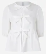 Blusa blanca de tricolina con cuello redondo, manga corta abullonada, cierre frontal con lazos y volado en la cintura.