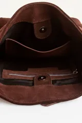 Cartera tipo balde de gamuza color marrón, con correa ajustable decorada con tachas metálicas plateadas y logo grabado en la parte frontal.