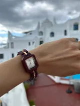 Reloj de pulsera con caja cuadrada y correa de eslabones de madera color borgoña. El dial es plateado con agujas y detalles dorados.