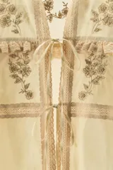Blusa color crema de algodón y viscosa, con cuello mao y mangas largas. Presenta bordados florales con abalorios y terminaciones en puntilla. Se cierra con lazos en el frente.