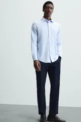 Camisa celeste a rayas verticales blancas, de corte recto, con cuello italiano abierto, mangas largas con puños abotonados y cierre frontal con botones.