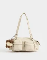 Cartera bandolera color crema con múltiples bolsillos frontales y laterales, doble asa corta regulable y charm de oso de peluche.