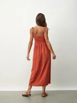 Vestido midi de color naranja, con escote recto y tirantes finos que se anudan sobre los hombros. Posee una cintura entallada con frunces que dan paso a una falda fluida.