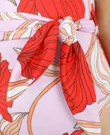 Traje de baño enterizo con estampado de flores rojas y hojas color crema sobre fondo lila. Presenta corte alto en la pierna y cinturón lila con lazada lateral.