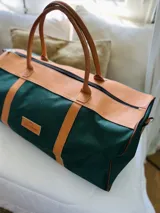 Bolso de lona verde oscuro con detalles en cuero color suela. Tiene doble asa de mano y cierre superior con cremallera.