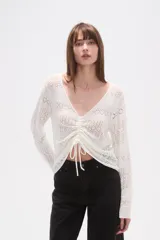 Sweater blanco de poliéster con cuello en V, manga larga y frunces ajustables en el frente. Presenta un tejido calado con patrones geométricos.