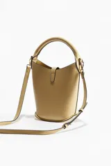 Cartera tipo bucket color beige, con asa de mano tubular y correa cruzada ajustable y extraíble. Cierre con trabilla y adorno metálico dorado en el frente. Forrada.