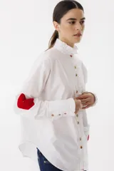 Camisa blanca de algodón con cuello alto con volados, cierre frontal con botones, mangas largas con puños abotonados, bolsillo en el pecho con corazón bordado y parches de crochet en los codos.