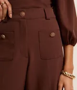 Pantalón sastrero marrón de corte recto y cintura alta, con trabillas en la cintura, bolsillos delanteros y traseros simulados, cierre tradicional y botones metalizados.