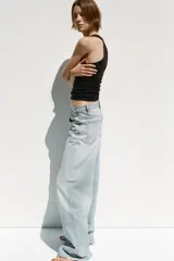 Pantalón de jean celeste de tiro alto, corte wide leg y largo hasta el tobillo. Presenta un diseño asimétrico en la cintura con cierre frontal cruzado con cremallera y botones metálicos.