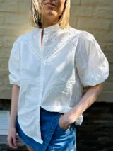 Camisa blanca de popelina de algodón, manga corta abullonada y cuello mao. Presenta doble detalle de puntilla en forma de V en la parte superior delantera.