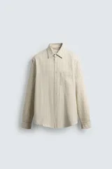 Camisa de hombre relaxed fit confeccionada en tejido de algodón a rayas verticales color beige y marrón. Tiene cuello solapa, manga larga con puños abotonados, bolsillo de parche en el pecho y cierre frontal con botones.