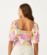 Top cropped de tejido tipo laise con estampado floral pixelado en tonos pastel (rosa, amarillo, azul, blanco). Presenta escote corazón con abertura central y lazo ajustable, y mangas cortas abullonadas.