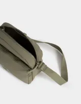 Bandolera gris claro de tela deportiva con bolsillo frontal, correa larga regulable, acceso con cierre superior y tag de goma.
