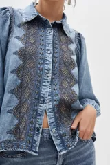 Camisa de denim celeste con bordados geométricos en el frente, corte recto, cuello camisero, manga larga con frunces en los hombros y puños abotonados.