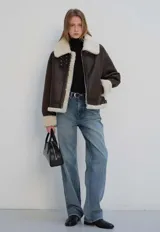 Chaqueta marrón de cuero vegano con forro de sherpa color crema. Presenta solapas con hebillas, cuello italiano, cierre de cremallera y mangas con puños.
