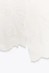 Blusa blanca de algodón con cuello solapa, escote pico y manga larga acampanada. Detalle de frunces y bordados a tono.