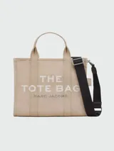Bolso tote mediano color beige con estampado frontal del logo "The Tote Bag Marc Jacobs" en blanco. Tiene doble asa corta y correa larga extraíble de tela azul marino.