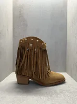 Bota corta de gamuza color marrón con flecos, tachas y apliques metálicos. Tiene taco bajo de madera.