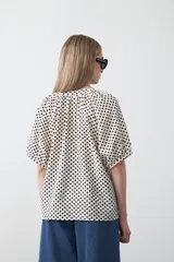 Camisa de viscosa con estampado de pequeños lunares verdes sobre fondo blanco o crema. Presenta escote en V con botones frontales y mangas cortas abullonadas con elástico en el puño.