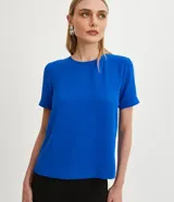 Blusa azul de poliéster con cuello redondo, mangas cortas con pespuntes, cierre trasero en forma de gota con botón y pequeñas aberturas laterales.