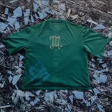 Remera verde oscuro de corte boxy/oversize con logo "M" bordado en dorado en la parte superior de la espalda.