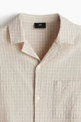 Camisa de manga corta color crema en tejido sirsaca de algodón. Modelo boxy con cuello abierto, tapeta lisa, bolsillo superior y bajo recto. Corte holgado.