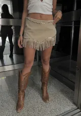 Minifalda color beige de gamuza sintética, con tiro bajo, corte irregular, tachas plateadas y flecos en el ruedo.
