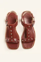 Sandalia de cuero color marrón con tiras finas, detalles de tachas metálicas y hebilla lateral.