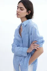 Camisa celeste y blanca a rayas verticales, de lino liviano, con cuello clásico, botones frontales, canesú de doble capa en la espalda, hombros caídos, mangas largas con puños abotonados, bolsillo de parche en el pecho y bajo redondeado.