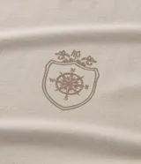 Remera de algodón color beige claro, manga corta y cuello redondo, con un pequeño estampado localizado en el pecho izquierdo de un escudo con una rosa de los vientos.