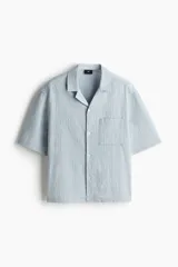Camisa celeste de manga corta en sirsaca de algodón. Modelo boxy con cuello abierto, tapeta lisa, bolsillo superior y bajo recto. Corte holgado.