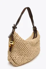Bolso tipo shopper color beige, con cuerpo trenzado de rafia y asa de hombro de cuero marrón. Tiene un charm colgante en un lateral y cierre con cremallera.